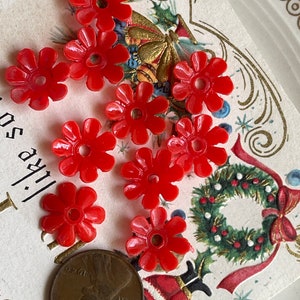 May include: A collection of ten red plastic flower-shaped beads. The beads have a small hole in the center and are arranged on a white surface with a Christmas card in the background.
