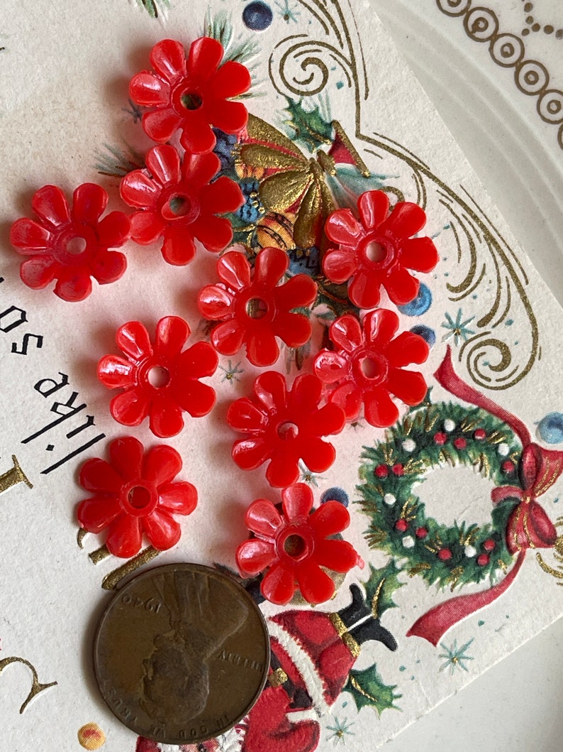 May include: A collection of ten red plastic flower-shaped beads. The beads have a small hole in the center and are arranged on a white surface with a Christmas card in the background.