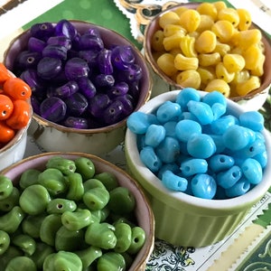 May include: Five small bowls filled with colorful glass beads. The bowls are white with gold trim. The beads are purple, yellow, blue, green, and orange.