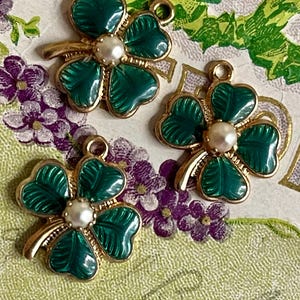 Puede incluir: Tres colgantes dorados con forma de tr&eacute;bol de cuatro hojas, con hojas de esmalte verde y una perla central. Cada colgante tiene un lazo para sujetarlo a joyas u otras manualidades. Los colgantes se muestran sobre un fondo con motivos florales.