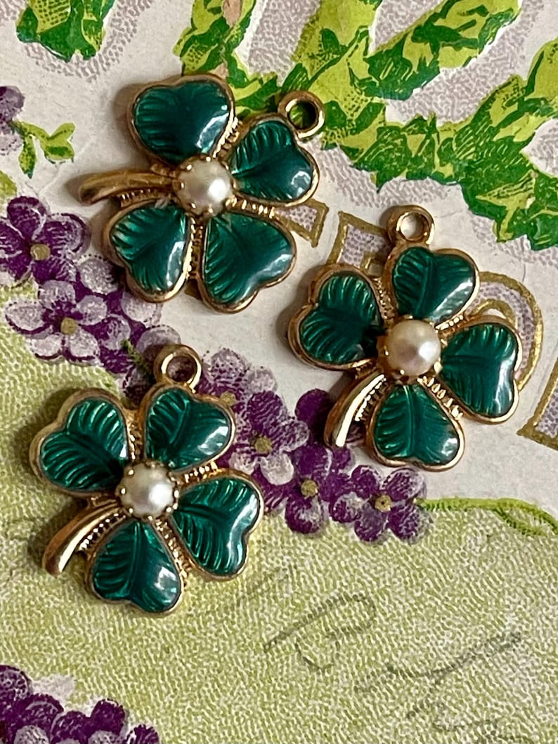 Puede incluir: Tres colgantes dorados con forma de tr&eacute;bol de cuatro hojas, con hojas de esmalte verde y una perla central. Cada colgante tiene un lazo para sujetarlo a joyas u otras manualidades. Los colgantes se muestran sobre un fondo con motivos florales.
