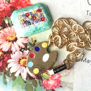 K&ouml;nnte beinhalten: Eine goldene Kettenkette mit einem goldenen Farbpaletten-Anh&auml;nger und einem kleinen quadratischen Anh&auml;nger in Gr&uuml;n mit einem Mosaik aus bunten Blumen. Die Kette ist im Seil-Stil und der Anh&auml;nger ist mit einem goldenen Verschluss befestigt. Die Kette liegt auf einer wei&szlig;en Oberfl&auml;che mit Text im Hintergrund.