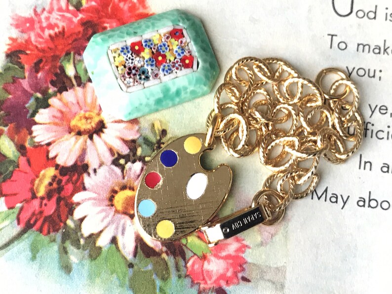 K&ouml;nnte beinhalten: Eine goldene Kettenkette mit einem goldenen Farbpaletten-Anh&auml;nger und einem kleinen quadratischen Anh&auml;nger in Gr&uuml;n mit einem Mosaik aus bunten Blumen. Die Kette ist im Seil-Stil und der Anh&auml;nger ist mit einem goldenen Verschluss befestigt. Die Kette liegt auf einer wei&szlig;en Oberfl&auml;che mit Text im Hintergrund.
