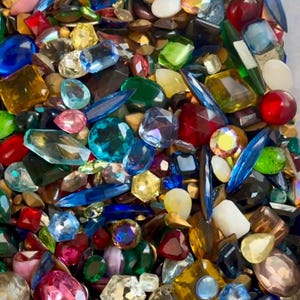 May include: A vibrant assortment of colorful gemstones in various shapes and sizes. The collection includes clear, blue, red, green, and pink stones, with faceted and smooth surfaces. The gemstones are scattered, creating a dazzling display.