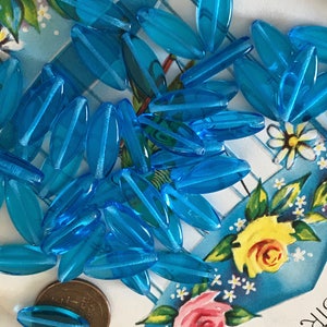 May include: A pile of turquoise blue glass beads in an oval shape. The beads are scattered on a blue and white background with flowers.
