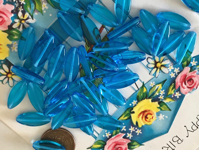 May include: A pile of turquoise blue glass beads in an oval shape. The beads are scattered on a blue and white background with flowers.