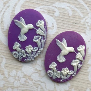 May include: Two purple oval-shaped cabochons with white raised designs of hummingbirds and flowers.
