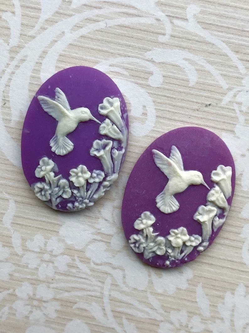 May include: Two purple oval-shaped cabochons with white raised designs of hummingbirds and flowers.