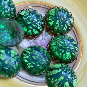 May include: A collection of seven green glass cabochons with gold foil accents. The cabochons are round and have a textured surface. They are arranged in a circle on a white plate with gold trim.