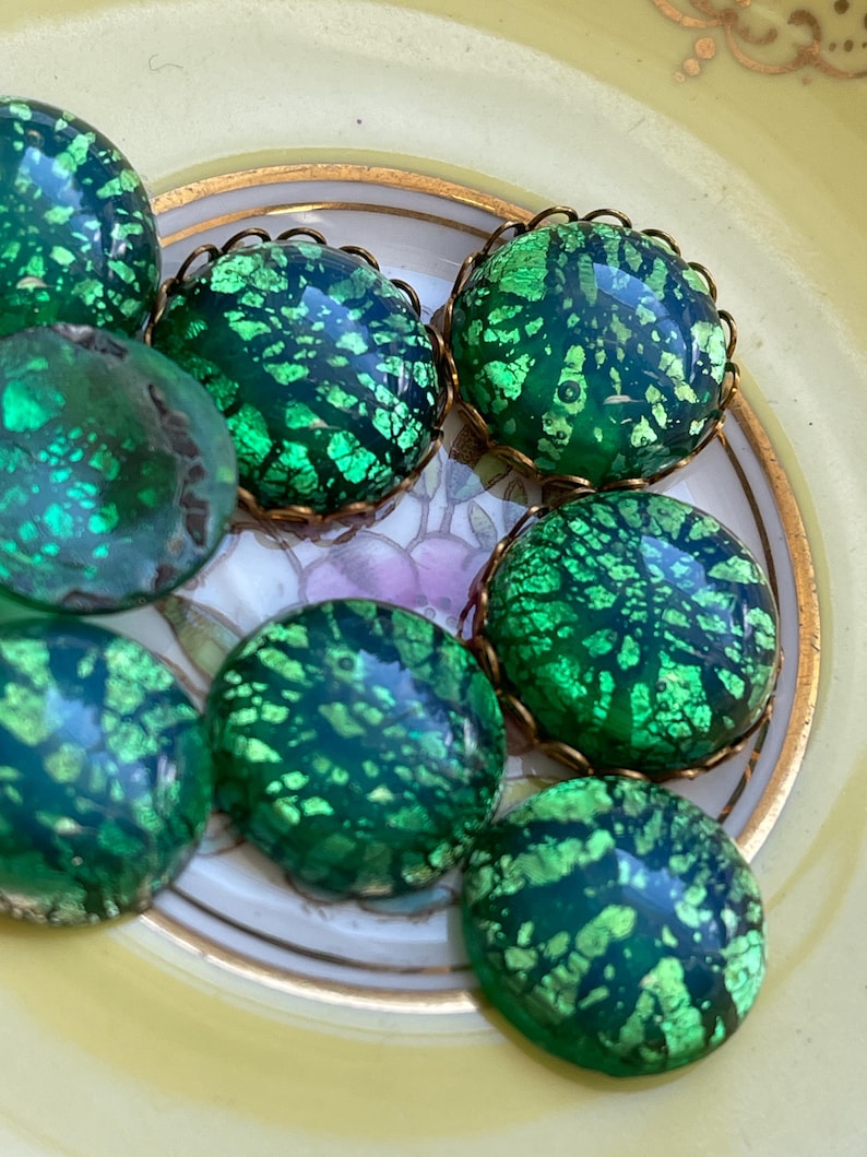 May include: A collection of seven green glass cabochons with gold foil accents. The cabochons are round and have a textured surface. They are arranged in a circle on a white plate with gold trim.
