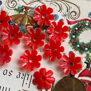 May include: A collection of ten red plastic flower-shaped buttons with a hole in the center. The buttons are arranged on a white surface with a Christmas-themed background.