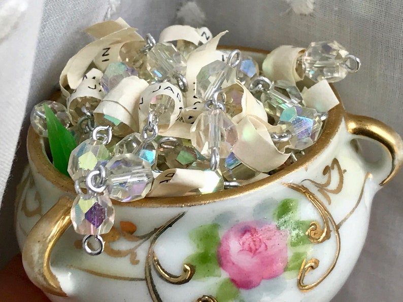 Puede incluir: Una tetera de porcelana blanca con ribetes dorados y un dise&ntilde;o de rosa rosa, llena de cuentas de vidrio iridiscentes y trozos de papel.
