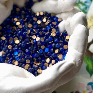 May include: A close-up of a pile of small, round, blue glass beads with gold accents. The beads are held in the cupped hands of a white ceramic sculpture.
