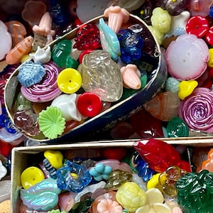 May include: A collection of colorful glass beads in various shapes and sizes, including flowers, leaves, and geometric shapes. The beads are arranged in two boxes, one with a blue lid and the other with a brown lid.