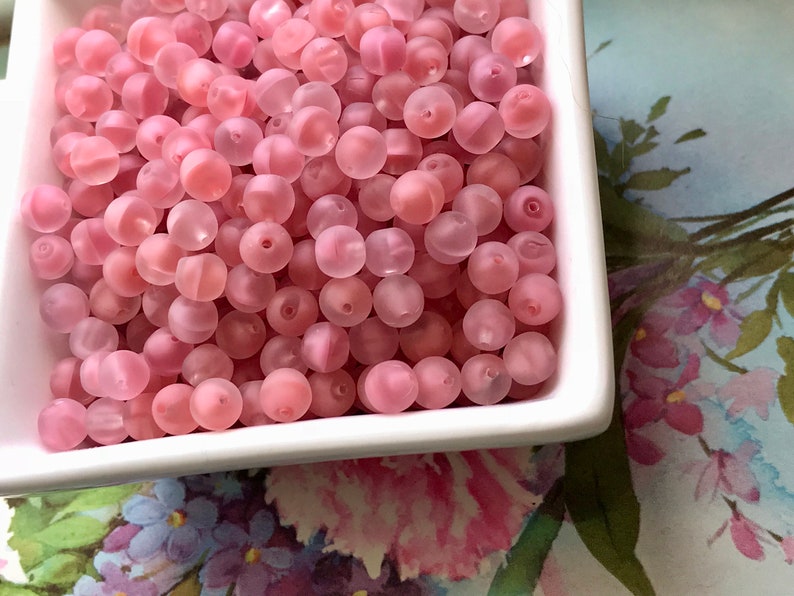 May include: A bowl filled with pink translucent round beads. The beads have a frosted finish and are perfect for jewelry making or crafting.