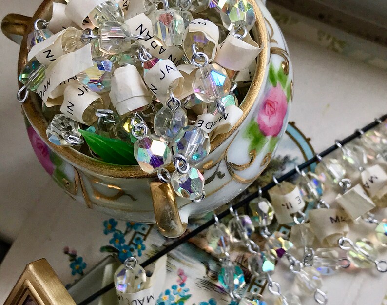 Puede incluir: Un taz&oacute;n de porcelana blanca con un dise&ntilde;o de rosa rosa, lleno de una hebra de cuentas de cristal iridiscente y peque&ntilde;as etiquetas de papel que dicen "Hecho en Jap&oacute;n".