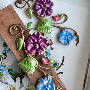 May include: Three metal flower charms with gold accents. The flowers are purple, blue, and pink. The flowers are attached to a gold stem with green leaves.
