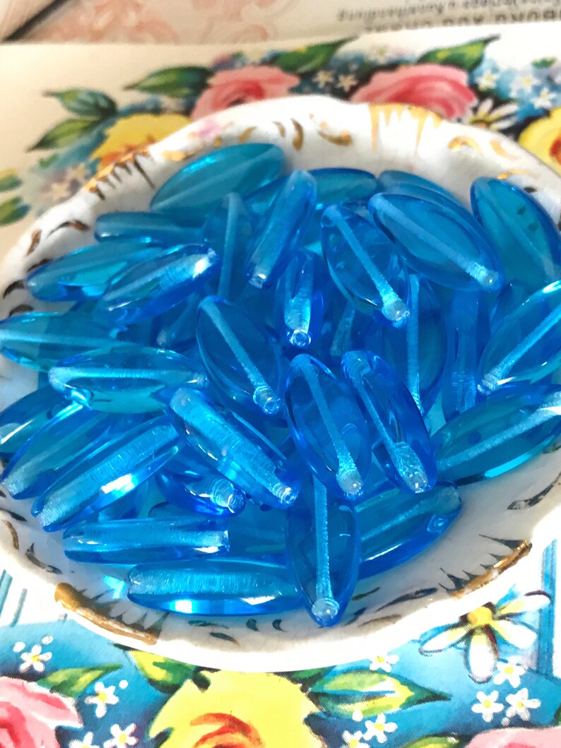 May include: A bowl filled with turquoise blue glass beads. The beads are oval shaped and have a smooth, glossy finish.