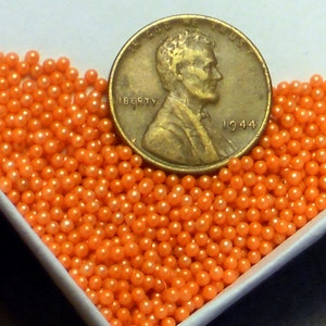 May include: A close-up of a white bowl filled with orange pearl sprinkles. A 1944 US penny is placed on top of the sprinkles for scale.