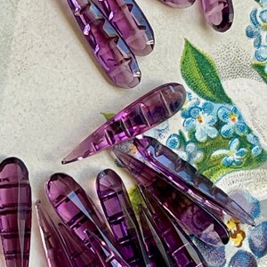 May include: A group of purple glass teardrop-shaped beads with a faceted design. The beads are arranged in a random pattern on a white surface with a floral design.
