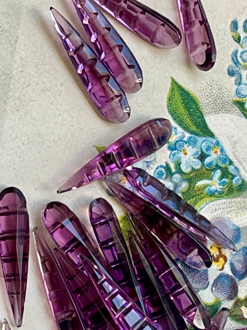 May include: A group of purple glass teardrop-shaped beads with a faceted design. The beads are arranged in a random pattern on a white surface with a floral design.