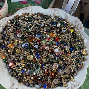 May include: A white decorative bowl filled with a collection of vintage jewellery pieces. The jewellery includes colourful rhinestones, gems, and metal settings in various shapes and sizes. The bowl sits on a green floral patterned surface.