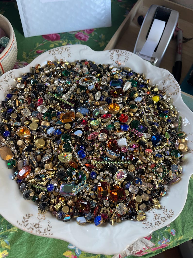 May include: A white decorative bowl filled with a collection of vintage jewellery pieces. The jewellery includes colourful rhinestones, gems, and metal settings in various shapes and sizes. The bowl sits on a green floral patterned surface.