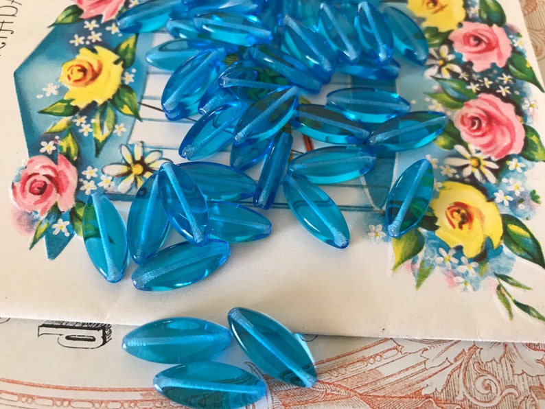 May include: A pile of light blue glass beads, oval shaped with a white stripe down the center. The beads are scattered on a white surface with a floral pattern.