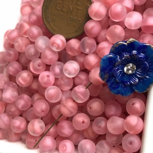May include: A bowl filled with pink frosted glass beads. The beads are round and have a slightly iridescent finish. A blue glass flower with a rhinestone center is sitting on top of the beads.