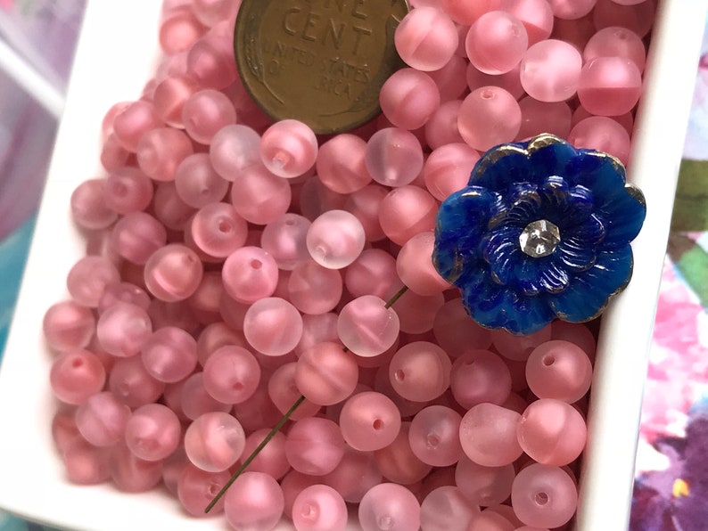 May include: A bowl filled with pink frosted glass beads. The beads are round and have a slightly iridescent finish. A blue glass flower with a rhinestone center is sitting on top of the beads.