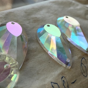 May include: Four iridescent teardrop-shaped glass beads with a hole at the top. The beads have a rainbow effect and are on a brown surface.