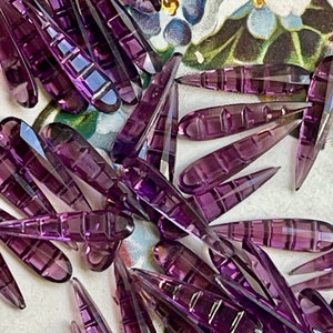 May include: A collection of purple glass teardrop-shaped beads with a faceted design. The beads are scattered on a floral patterned surface.