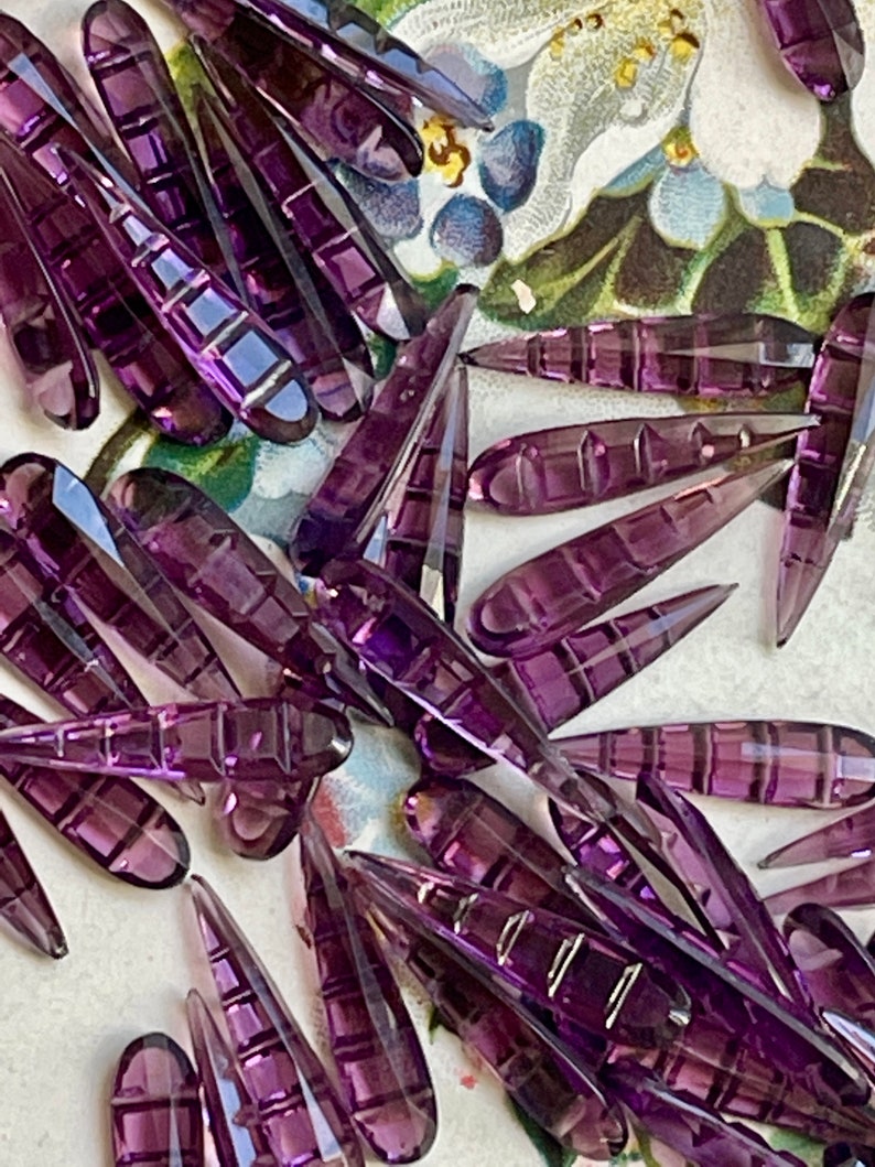 May include: A collection of purple glass teardrop-shaped beads with a faceted design. The beads are scattered on a floral patterned surface.