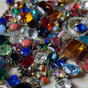 May include: A selection of colourful vintage rhinestone and glass jewels in various shapes and sizes. The assortment includes clear, red, blue, green, and yellow stones, some with gold-tone metal settings. A variety of faceted and cabochon cuts are visible.