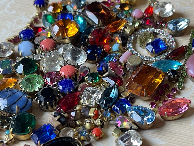 May include: A selection of colourful vintage rhinestone and glass jewels in various shapes and sizes. The assortment includes clear, red, blue, green, and yellow stones, some with gold-tone metal settings. A variety of faceted and cabochon cuts are visible.
