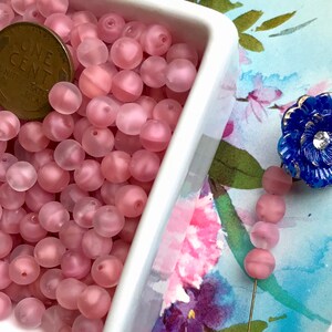 May include: A white bowl filled with pink frosted glass beads. The beads are round and have a slightly iridescent finish. A penny is included for scale.