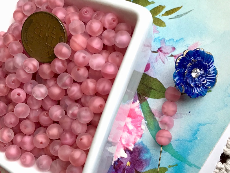 May include: A white bowl filled with pink frosted glass beads. The beads are round and have a slightly iridescent finish. A penny is included for scale.