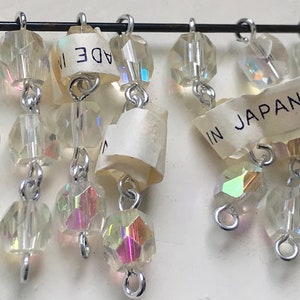 Puede incluir: Una hebra de cuentas de vidrio transparente iridiscente con bucles de metal plateado, colgando de un alambre plateado. Las cuentas est&aacute;n enhebradas en un patr&oacute;n repetitivo de tres cuentas, luego un bucle, luego tres cuentas. La hebra est&aacute; etiquetada como "MADE IN JAPAN".