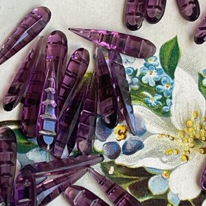 May include: A collection of purple glass teardrop-shaped beads with a faceted design. The beads are scattered on a floral patterned surface.
