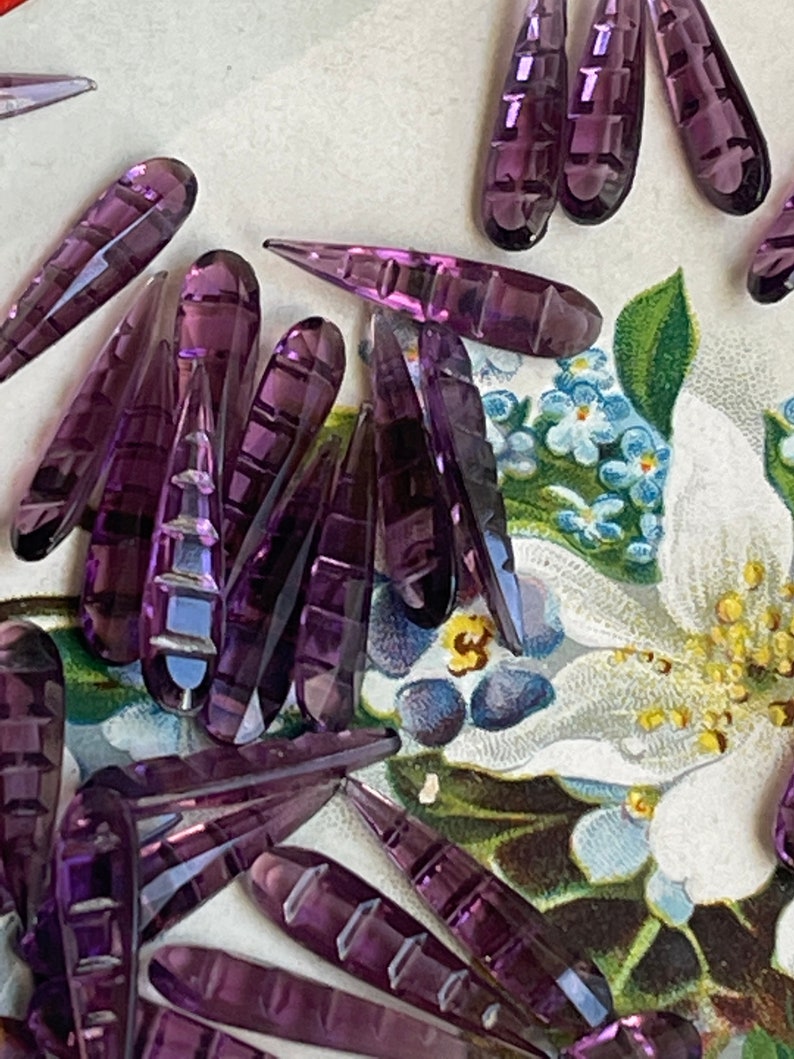 May include: A collection of purple glass teardrop-shaped beads with a faceted design. The beads are scattered on a floral patterned surface.