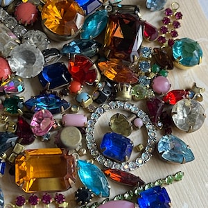May include: A selection of vintage rhinestones and jewels in a variety of colours and forms. The collection features clear, blue, red, yellow, and pink stones, some set in metal. A sparkling, circular rhinestone piece is also included.