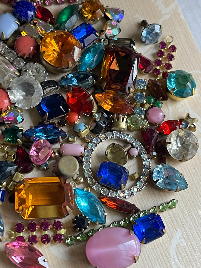 May include: A selection of vintage rhinestones and jewels in a variety of colours and forms. The collection features clear, blue, red, yellow, and pink stones, some set in metal. A sparkling, circular rhinestone piece is also included.