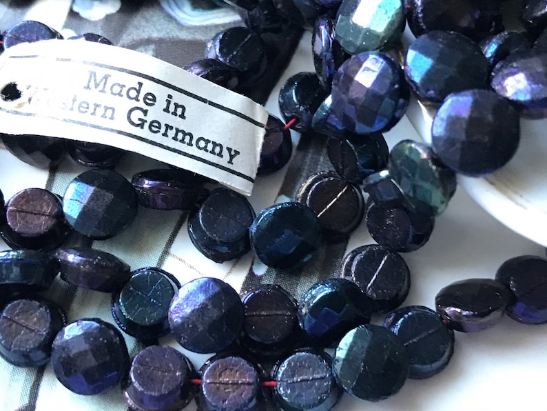 Puede incluir: Una hebra de cuentas de vidrio azul oscuro iridiscente con una etiqueta que dice "Made in Western Germany".