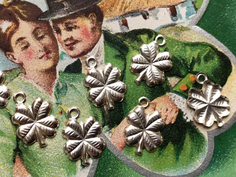 May include: Six silver four-leaf clover charms with a loop at the top for attaching to a necklace or bracelet. The charms are on a green and white background.