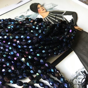May include: A strand of dark blue iridescent glass beads. The beads are faceted and have a slightly oval shape.