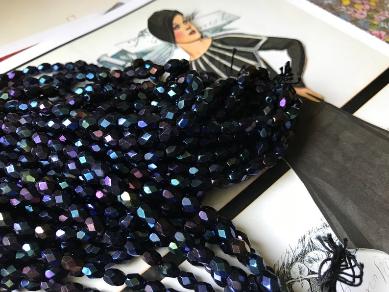 May include: A strand of dark blue iridescent glass beads. The beads are faceted and have a slightly oval shape.