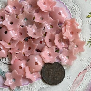 Peut inclure: Un ensemble de boutons en plastique rose en forme de fleur avec un trou au centre. Les boutons sont &eacute;parpill&eacute;s sur une surface blanche avec un motif de dentelle.