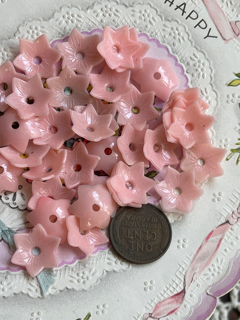 Peut inclure: Un ensemble de boutons en plastique rose en forme de fleur avec un trou au centre. Les boutons sont &eacute;parpill&eacute;s sur une surface blanche avec un motif de dentelle.