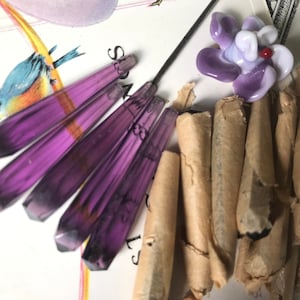 May include: An assortment of glass art pieces. The image shows several elongated, faceted purple glass pendants, a purple and white flower-shaped glass bead with a red centre, and several small, rolled paper items. The background includes a vintage illustration.