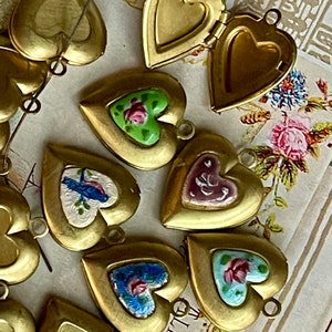 May include: A collection of heart-shaped locket pendants with various enamel designs. Some lockets feature floral patterns, while others have a bird or abstract design. The lockets are made of gold-toned metal and have a hinged opening.